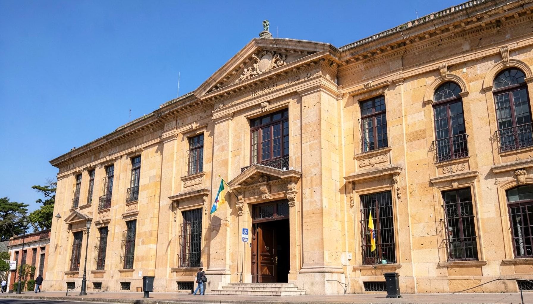A stately building facade in warm sandstone tones under a clear blue sky, with the Ecuadorian flag visible near the entrance, conveying diplomatic presence and formality.