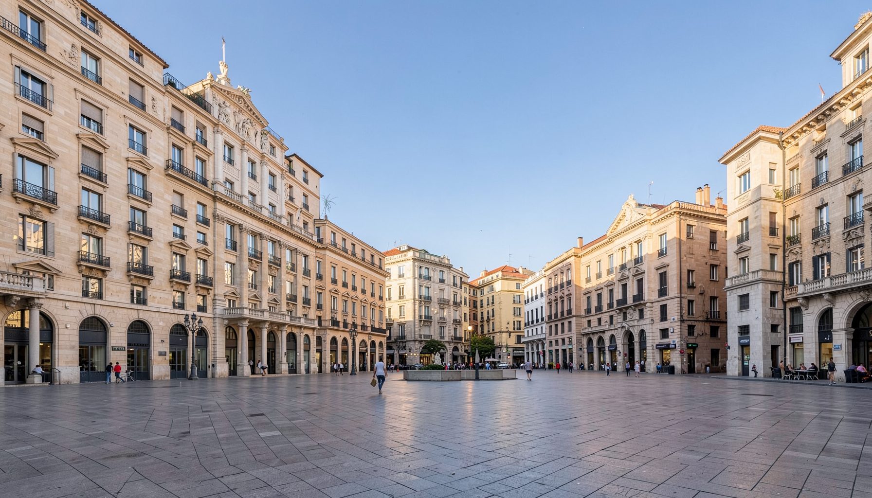 A wide-angle view of a modern city plaza in Madrid under soft afternoon light, with sandstone-colored buildings and a clear blue sky.