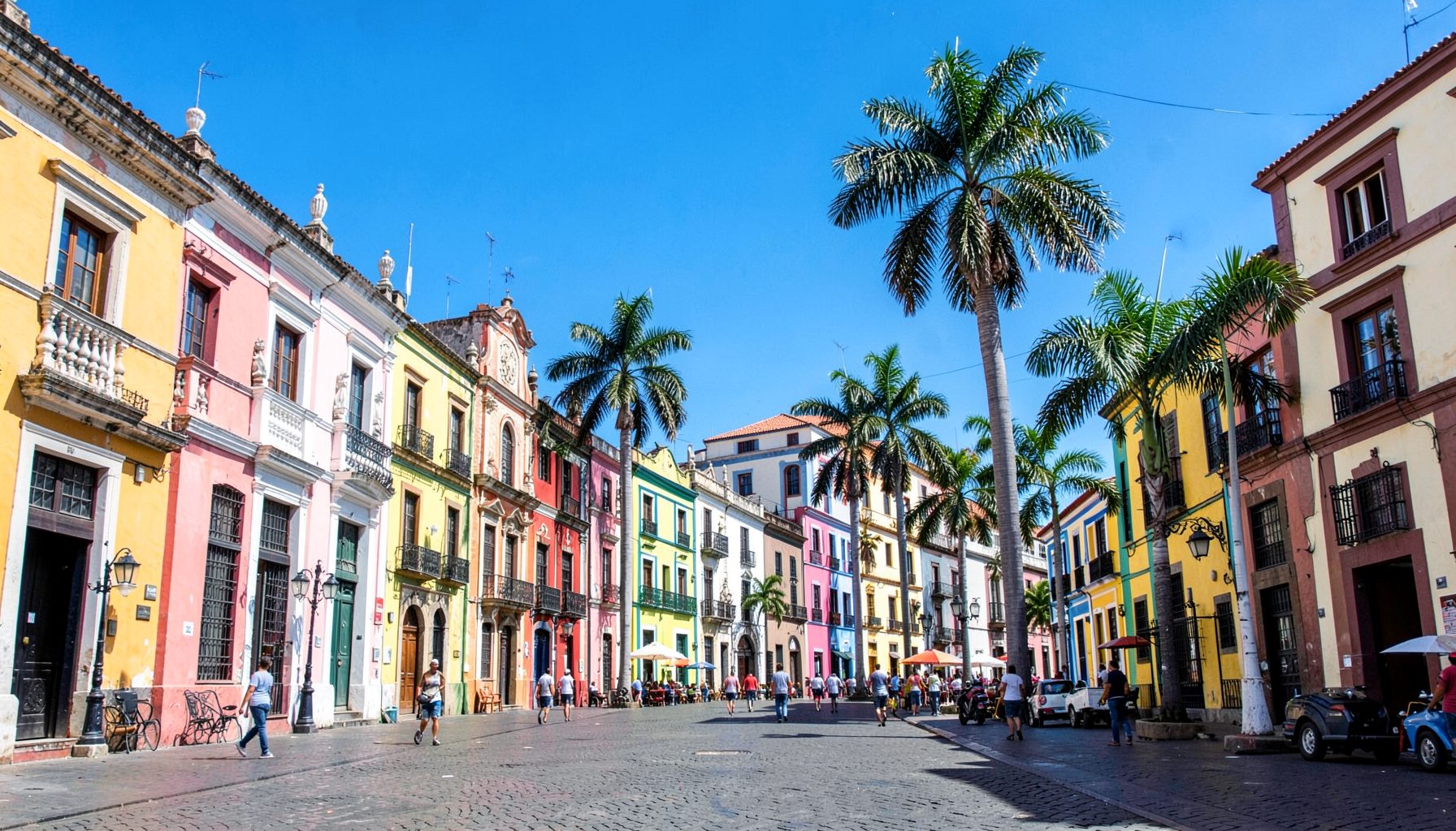 A vibrant street scene in a Latin American capital with colorful colonial architecture, clear blue sky, and palm trees lining a plaza